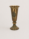 An antique-style bronze goblet or cup with a flared rim, decorative relief motif on the body, and a ribbed pedestal base, shown against a plain light background.