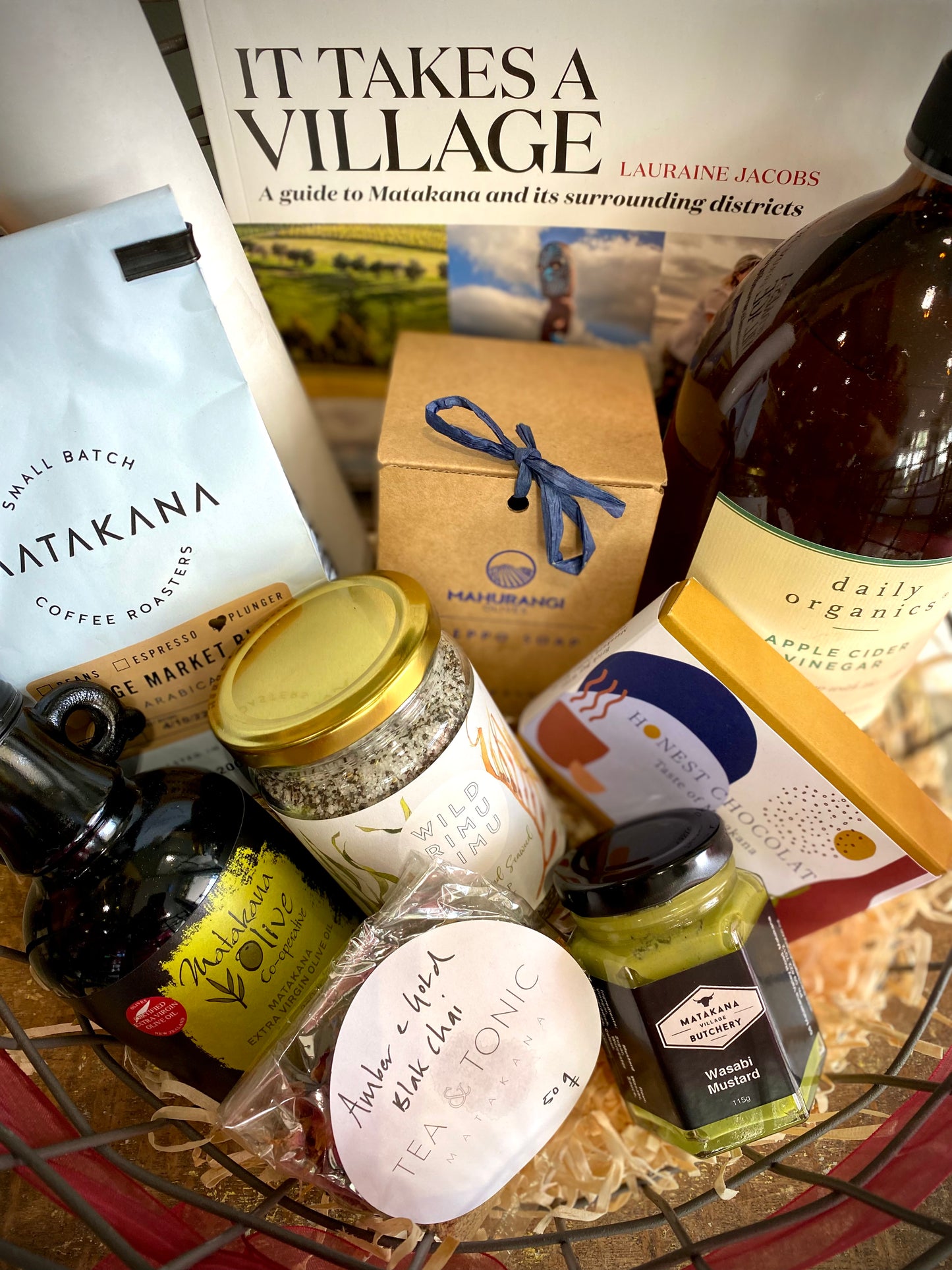 Close up of Wire hamper with Matakana olive oil, Matakana Coffee Roasters coffee, Daily Organics apple cider, Tea & Tonic Chai tea, Mahurangi Allepo soap, Matakana Butchers' mustard, Honest Chocolates, dried flowers, Wild Rimu Rimu salt and It Takes A Village book.